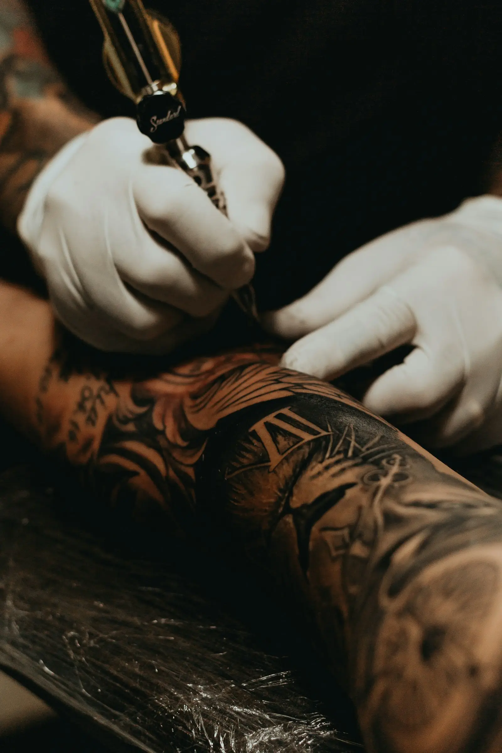 Mains gantées en train de tatouer un bras déjà couvert de tatouages, sous une lumière chaude